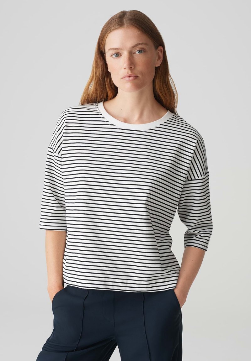Loose-fit, white top with black horizontal stripes, rounded neckline, and 3/4 sleeves. Made from a soft fabric with a smooth texture.
