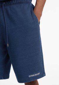 Mornarsko plave sportske kratke hlače od mekog materijala, s elastičnim pojasom i vezicom te bočnim džepovima. Uključuje logo "GUESS JEANS".