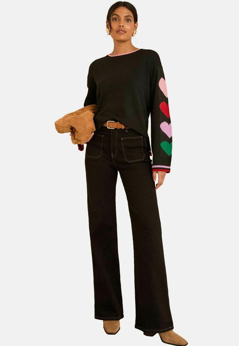 Schwarzer Pullover mit bunten Herzmustern an den Ärmeln, kombiniert mit hoch geschnittenen schwarzen Jeans und hält eine braune Wildlederjacke.