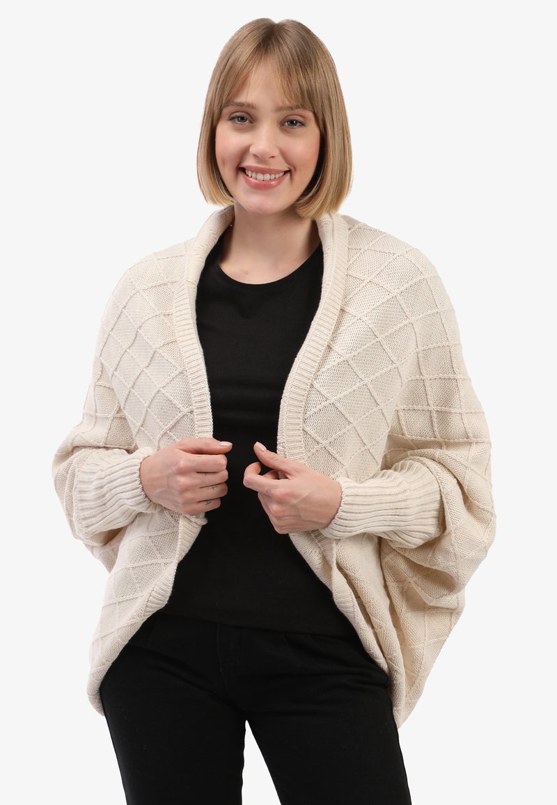 Lachende vrouw met kort blond haar, gekleed in een crèmekleurige gebreide cardigan over een zwart shirt en zwarte broek, poseert met haar handen die de cardigan vasthouden.
