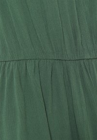 Zelená, splývavá tkanina s texturovaným povrchem a vertikálními záhyby; elastický pas viditelný v oblasti pasu.