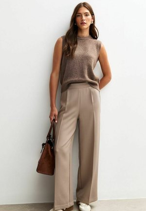 Brown sleeveless sweater paired with beige wide-leg trousers. Model holds a brown handbag. Textured fabric and relaxed fit.