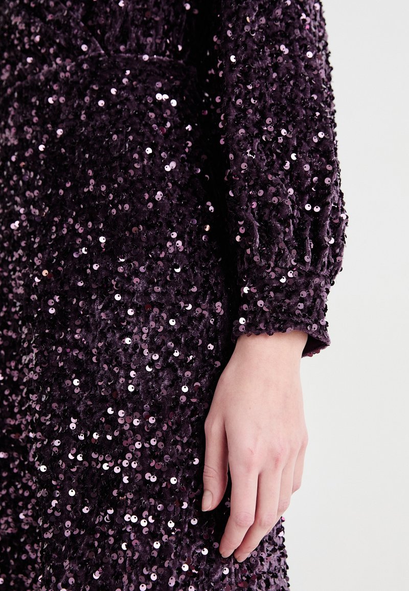 Fioletowa aksamitna tkanina ozdobiona błyszczącymi cekinami; szczegółowe długie rękawy z marszczonym nadgarstkiem; ręka częściowo widoczna, z neutralnym lakierem do paznokci.