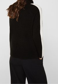 Kobieta w czarnym swetrze z białym paskiem na rękawie, stojąca z rękami w kieszeniach, zwrócona tyłem do kamery.