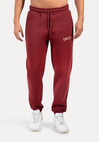Bordeauxkleurige joggingbroek met een elastische tailleband en trekkoord. Beschikt over een zak en een witte geborduurde logo op de bovenbeen. De stof is zacht en glad.