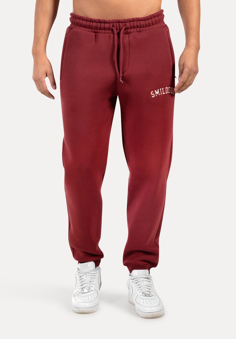 Bordeauxkleurige joggingbroek met een elastische tailleband en trekkoord. Beschikt over een zak en een witte geborduurde logo op de bovenbeen. De stof is zacht en glad.