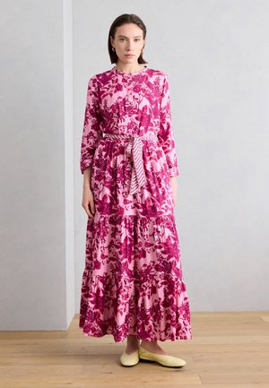 Mujer vestida con un vestido largo rosa con flores, un cinturón estampado y zapatos planos amarillos, de pie sobre un suelo de madera frente a una pared lisa.