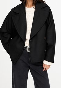 Sort dobbeltradet jakke med store revers, tekstureret stof, to knapper, båret over en cremefarvet strikket sweater, kombineret med mørke jeans.