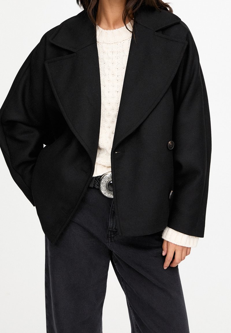 Sort dobbeltradet jakke med store revers, tekstureret stof, to knapper, båret over en cremefarvet strikket sweater, kombineret med mørke jeans.