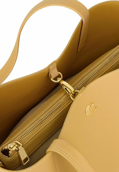 Bež usnjena torbica z zlatim zadrgo in zaponko, ki ima prostorno notranjost in majhno vrezano črko "C" na sprednji preklopni površini.