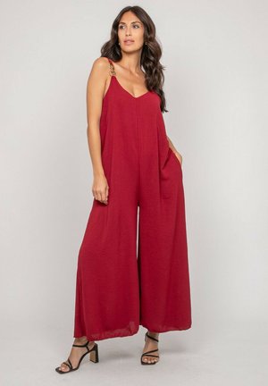 Femme portant une combinaison rouge sans manches à jambes larges avec des bretelles en chaîne, associée à des sandales noires à talons.