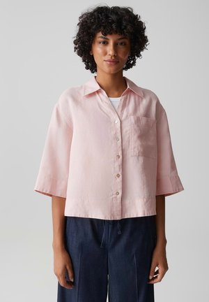 Frau mit lockigem Haar trägt ein lockeres, hellrosa Hemd mit Knöpfen und ellbogenlangen Ärmeln sowie dunkelblaue Hosen, steht vor einem einfachen Hintergrund.