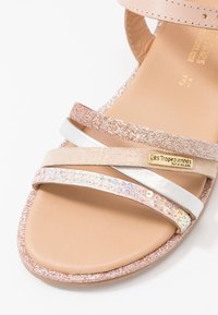 Sandalia con tiras y suela beige. Presenta un tirante brillante en oro rosa, plata y un tirante con lentejuelas, destacando varias texturas y acabados.