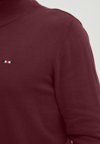 Burgundy villapaita korkealla kaulalla. Rintamuksessa pieni, erottuva logo, jossa on punaisia, valkoisia ja sinisiä raitoja. Pehmeä pinta.