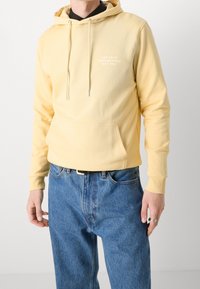 Homme portant un sweat à capuche jaune pâle avec poche devant, texte "Les Deux Copenhagen Est. 2011", associé à un jean bleu et une bague en argent à la main droite.