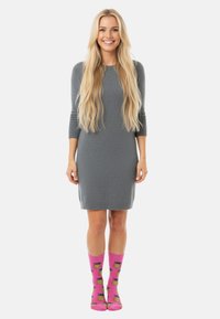 Mujer sonriente con cabello largo y rubio que lleva un vestido gris hasta la rodilla y calcetas rosas brillantes con patrones de piña amarilla.