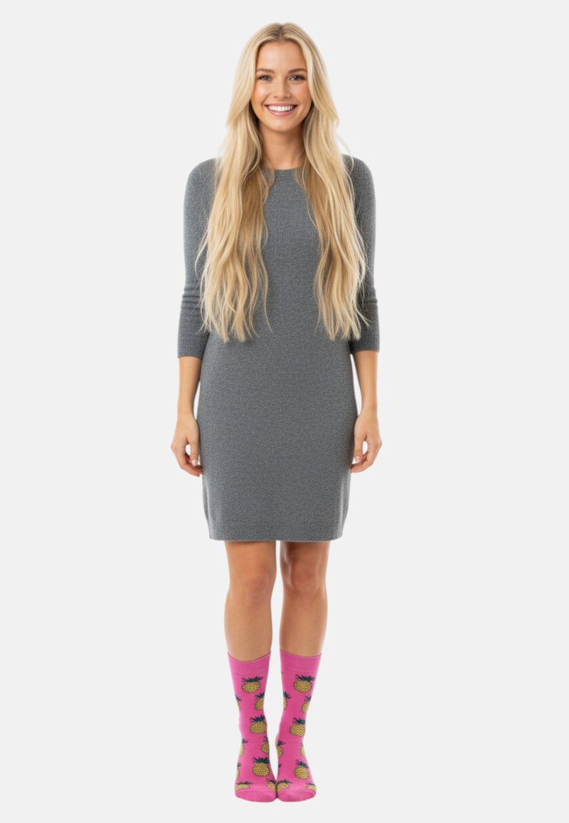 Mujer sonriente con cabello largo y rubio que lleva un vestido gris hasta la rodilla y calcetas rosas brillantes con patrones de piña amarilla.
