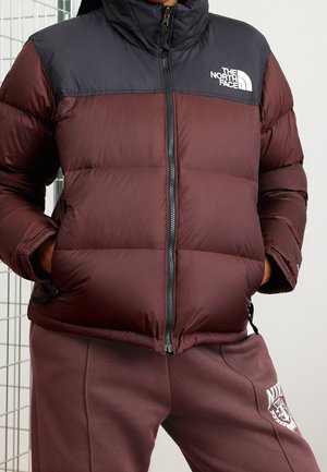 Personne portant une doudoune zippée bordeaux et noire avec le logo "The North Face" sur la poitrine, et un pantalon de survêtement bordeaux avec un emblème blanc.