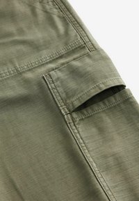 Gros plan sur un tissu vert olive avec une poche rectangulaire dotée d'un rabat et de détails de double surpiqûre visibles.