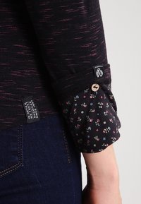 Dunkles Langarmhemd mit gesprenkeltem Muster, floraler Manschette mit Knopf, kombiniert mit dunkelblauen Jeans.