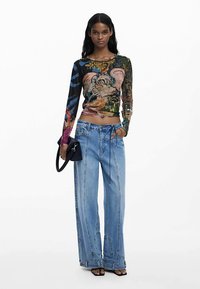 Langärmliges figurbetontes Oberteil mit buntem Blumen- und Tierprint, kombiniert mit lockeren hellblauen Jeans mit Steppnähten. Schwarze Handtasche.