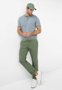 Polo shirt a strisce azzurro chiaro con dettagli verdi, abbinato a pantaloni a sigaretta verde oliva e sneakers bianche. Il modello è in piedi in modo casual contro un muro.