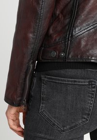 Braune Lederjacke mit matter Oberfläche, Reißverschluss am Ärmel und Druckknopfverschluss, kombiniert mit schwarzen Jeans aus Denim mit einer Gesäßtasche.