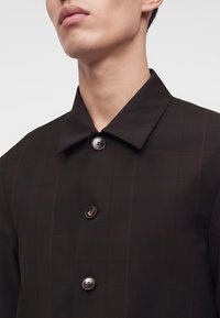Gros plan d'une personne portant un manteau sombre à boutons avec un col et trois boutons ronds visibles.