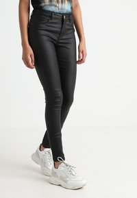 Woman wearing black slim-fit leather pants and white chunky sneakers, standing against a white background.