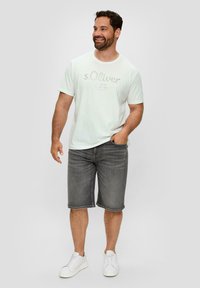 Biała koszulka krótki rękaw z jasnoszarym napisem, w połączeniu z szarymi szortami dżinsowymi i białymi trampkami. Model stoi w swobodnej postawie, z rękami w kieszeni.