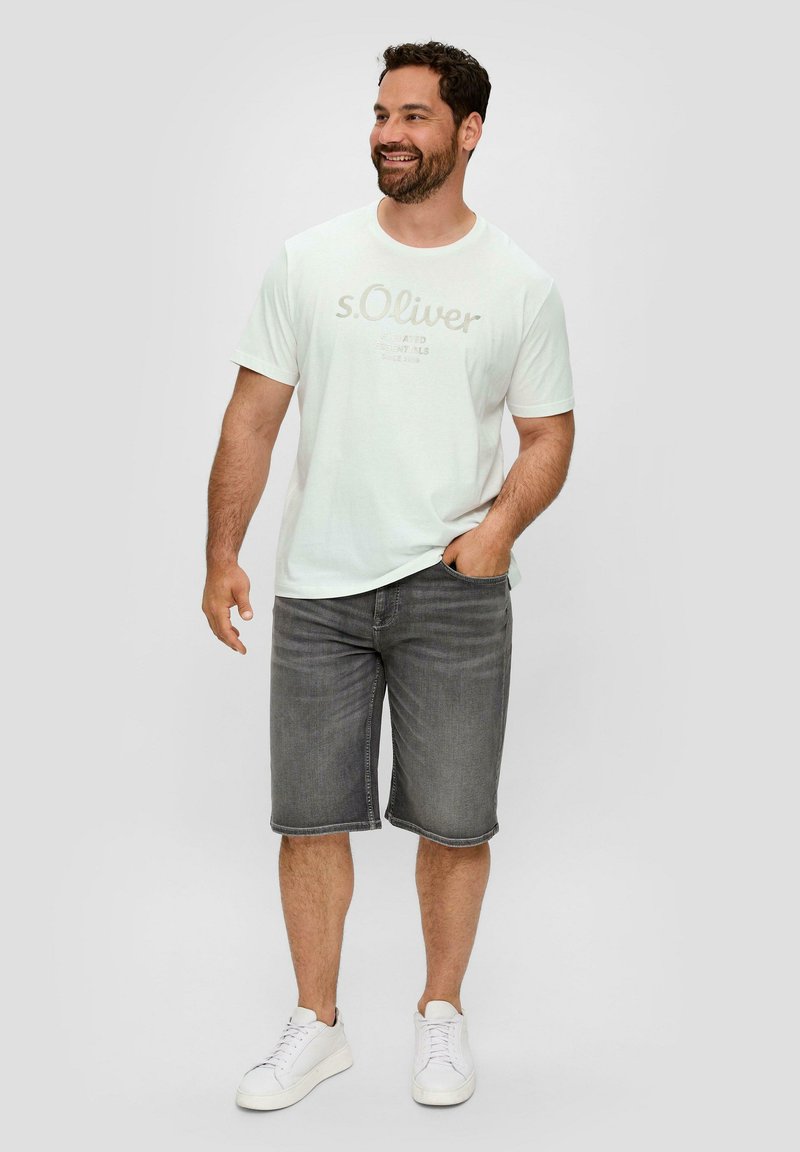 Biała koszulka krótki rękaw z jasnoszarym napisem, w połączeniu z szarymi szortami dżinsowymi i białymi trampkami. Model stoi w swobodnej postawie, z rękami w kieszeni.