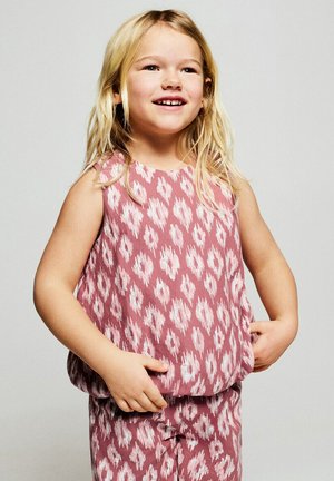 Jeune fille souriante aux longs cheveux blonds, portant un haut sans manches rose et blanc à motifs assortis à un pantalon, les mains reposant près des poches.