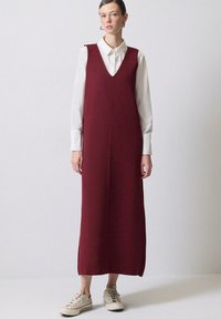 Burgundy ärmlös klänning med V-ringning och sidoflikar, matchad med en vit skjorta med krage och beige sneakers. Slät tygtextur.