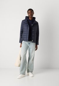 Giacca puffer blu navy con design trapuntato, chiusura con zip e cappuccio, abbinata a jeans a gamba larga blu chiaro e sneakers bianche. Borsa chiara in mano.