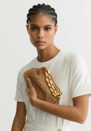Woman with braided hair in white shirt holds a woven brown clutch bag with a large gold chain accent.
