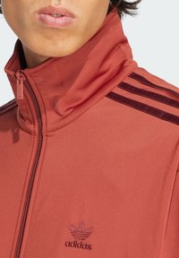 Chaqueta roja con cremallera y cuello alto, que presenta tres rayas oscuras en el hombro y un logo bordado de Adidas en el pecho.