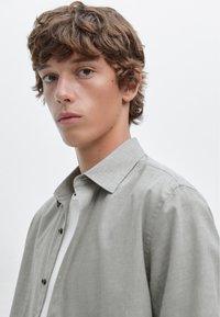 Lichtgrijze button-up shirt met een gestructureerde kraag, gedragen over een wit t-shirt. De stof is glad, met subtiele textuur en een minimaal ontwerp.