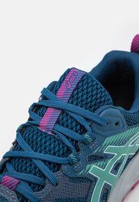 Niebieski but sportowy z siateczkową cholewą, zielonymi akcentami, różową zakładką z logo w paski i teksturowaną podeszwą. Bogata w funkcje konstrukcja zapewniająca oddychalność i wsparcie.