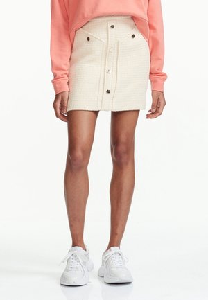 Weiße strukturierte Rock mit Vorderknöpfen und Taschen, kombiniert mit einem rosa Sweatshirt und weißen Sneakern. Fokus auf dem Stoff und den Knopfdetails.