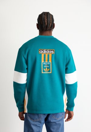 Mann mit geflochtenen Haaren, der einen türkisfarbenen Adidas-Sweatshirt mit weißen und gelben Streifen, Logo und Text auf der Rückseite trägt, kombiniert mit blauen Jeans.