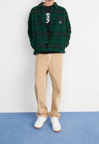 Camicia di flanella a quadri verde e nera, pantaloni beige e sneakers bianchi. La camicia presenta bottoni e un dettaglio sulla tasca frontale.