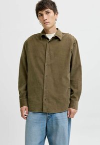 Giovane uomo con capelli corti castani, che indossa orecchini a cerchio argentati, una camicia bottonata in corduroy verde oliva e jeans larghi azzurri, su sfondo bianco.