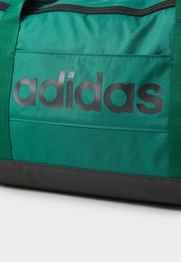 Grønn duffelbag med svart bunn og stor svart "adidas"-logo. Har solid stoff og strukturert overflate for holdbarhet.