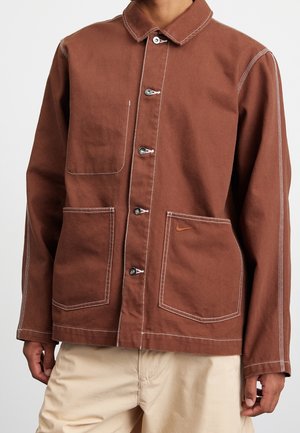 Mężczyzna ubrany w brązową kurtkę na guziki z dużymi kieszeniami, białym przeszyciem i małym pomarańczowym logo Nike na dolnej prawej kieszeni.