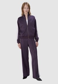 Fioletowa satynowa kurtka dresowa z białymi bocznymi paskami, zamkiem z przodu i pasującymi szerokimi spodniami. Gładka faktura i luźny krój.