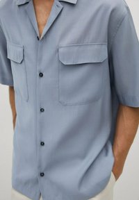Chemise bleu clair à manches courtes en tissu lisse, avec deux poches frontales, boutons noirs et coupe décontractée.