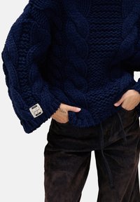 Granatowy sweter dziany z warkoczowymi wzorami, grubą fakturą i szerokimi rękawami. Na mankiecie małą metkę; zestawiony z ciemnobrązowymi spodniami.