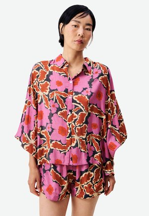 Femme portant une chemise ample à boutons et un short assorti avec de grands motifs floraux roses, orange et marron sur un fond clair.
