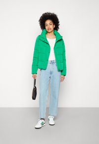 Lys grønn pufferjakke med høy krage, med knappestengning og quiltet design; kombinert med lyseblå rette jeans og hvite sneakers.