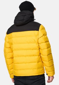 Gul puffjakke med en sort hætte og skulderområde, der har et quiltet design og elastiske manchetter. Bagfra ses en glat tekstur.